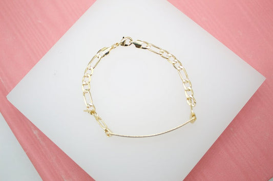 A delicate gold Figaro chain bracelet rests on a white display surface against a pink background.