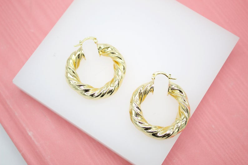 A pair of shiny gold twisted hoop earrings rest neatly on a white display block against a soft pink background.