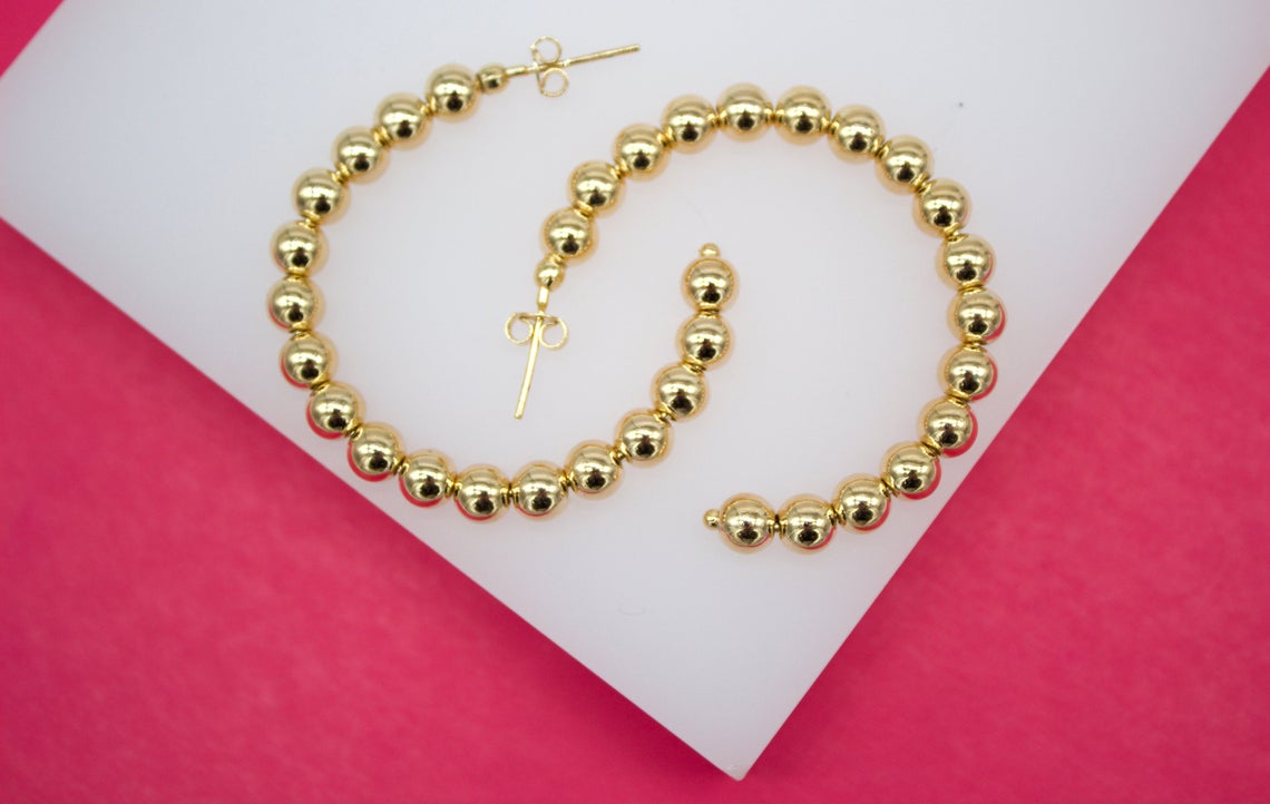 A pair of large, polished gold beaded hoop earrings rests on a white and pink surface.
