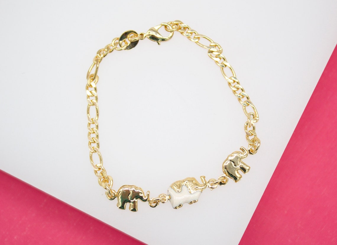A gold-plated link bracelet features three charming elephant charms displayed against a white and pink background.