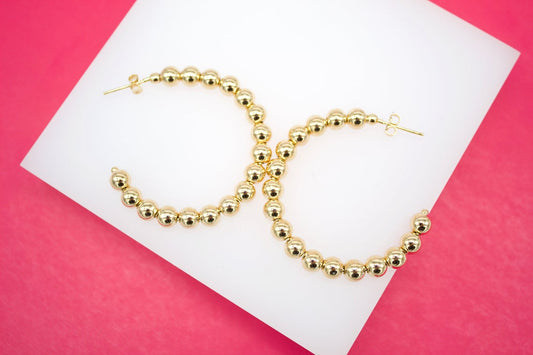 A pair of shiny gold ball hoop earrings rests on a white display against a vibrant pink background.