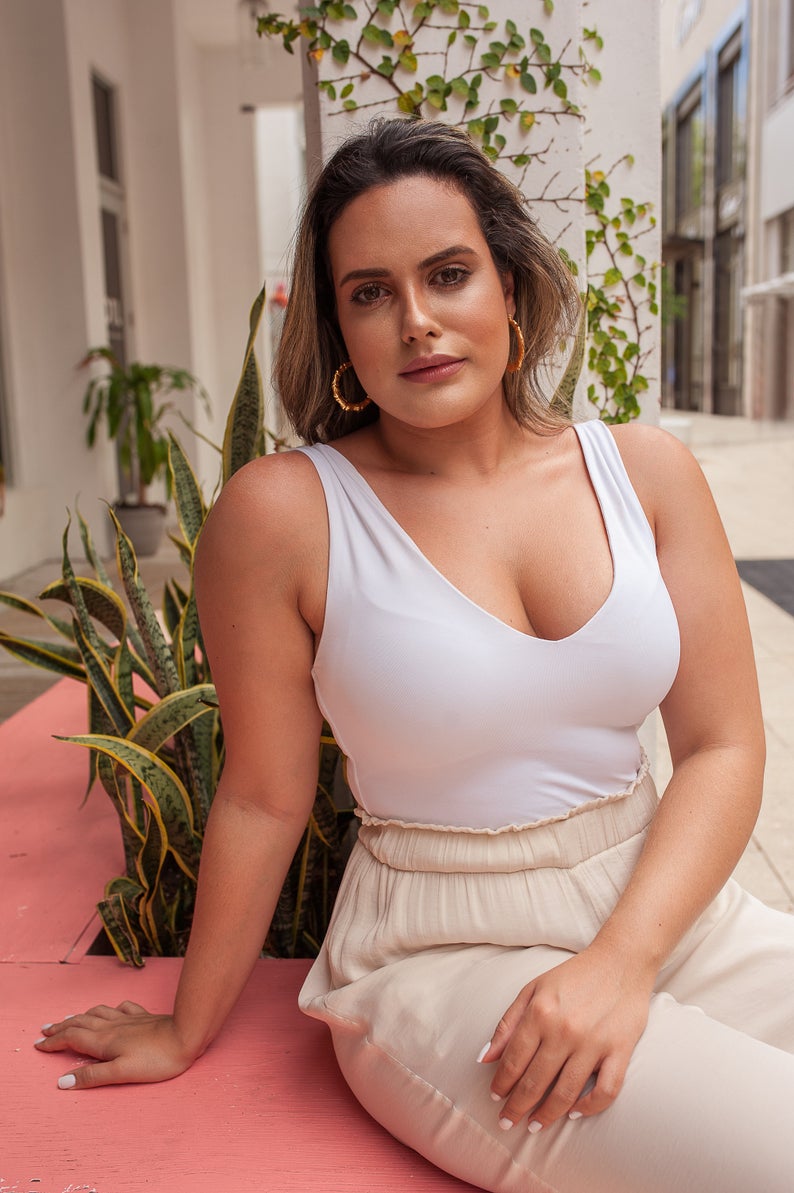 A plus-size brunette woman wearing a white tank top and beige pants relaxes outdoors near lush green plants.