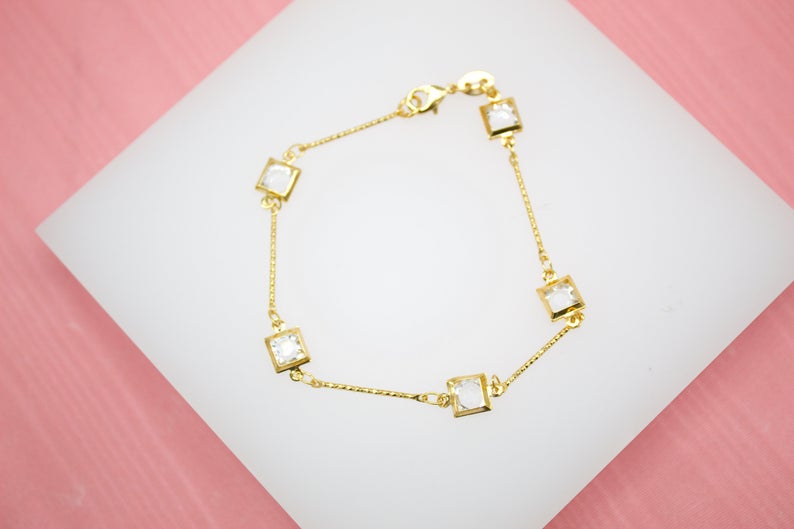A delicate gold chain bracelet features sparkling square white crystal charms arranged against a white and pink background.