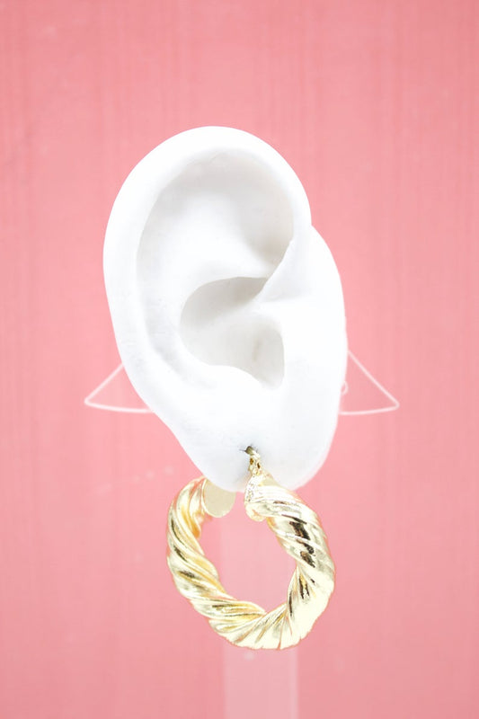 A white mannequin ear displays a chunky, twisted gold hoop earring against a soft pink background.