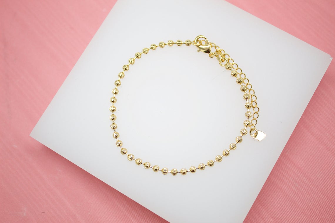 A delicate gold ball chain bracelet rests on a white display against a pink background.