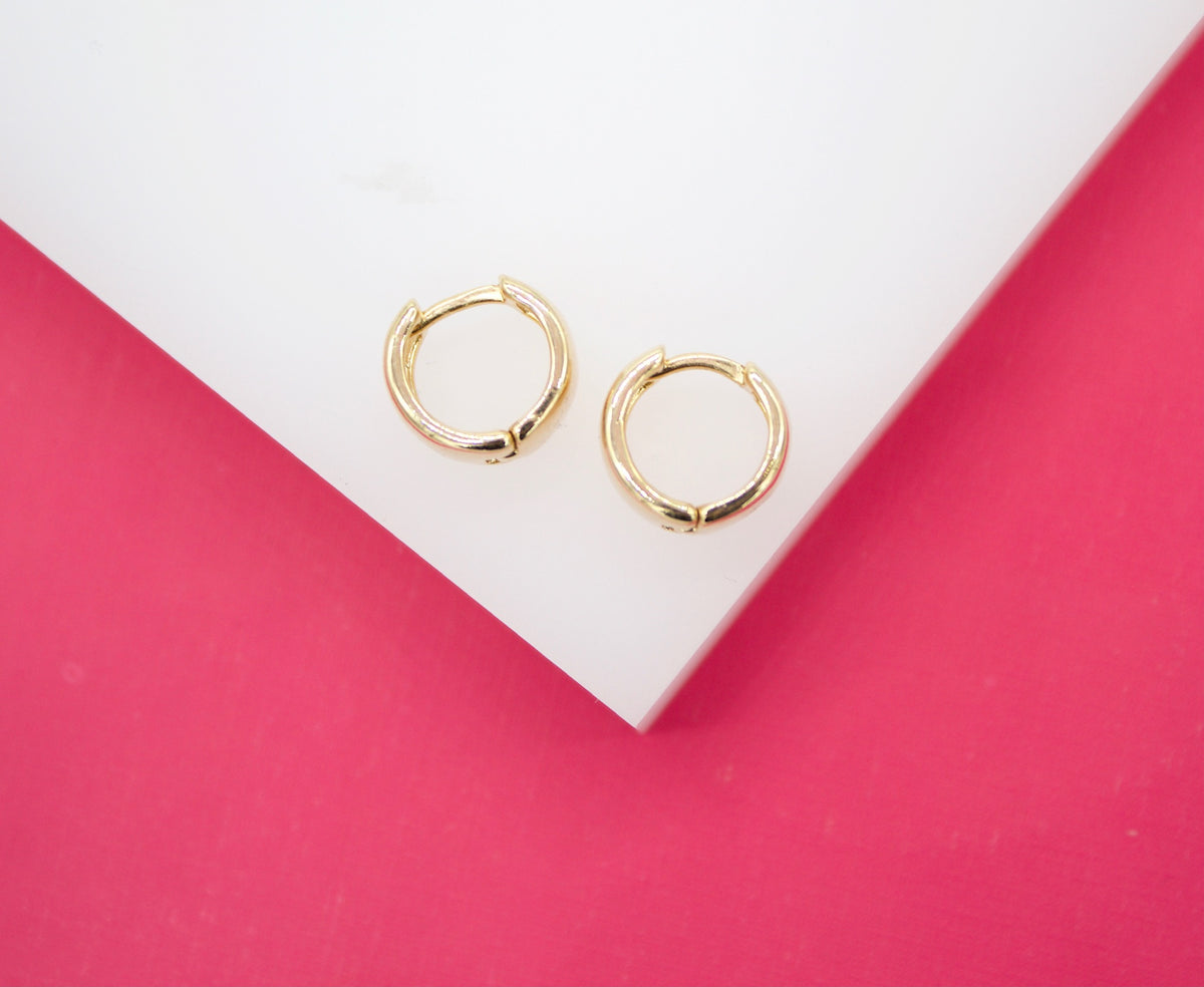 Two small gold hoop earrings rest on a white surface against a bright pink background.