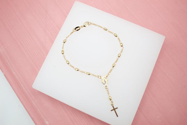 A delicate gold rosary bracelet rests on a white square display card against a pink textured background.