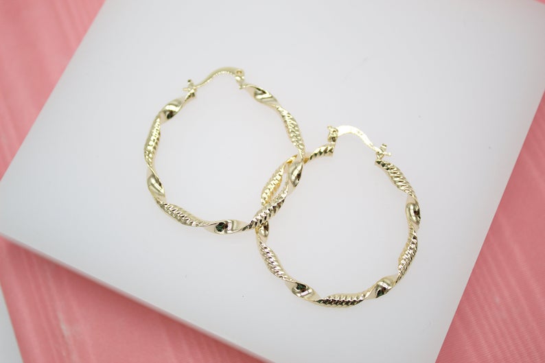 A pair of twisted gold hoop earrings rests on a white display surface against a pink background.