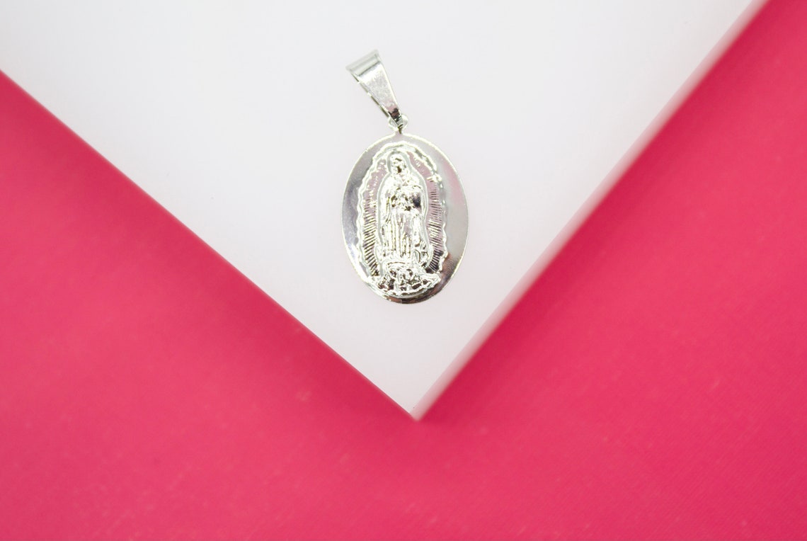 A silver oval Our Lady of Guadalupe pendant rests on a white geometric display against a vibrant pink background.