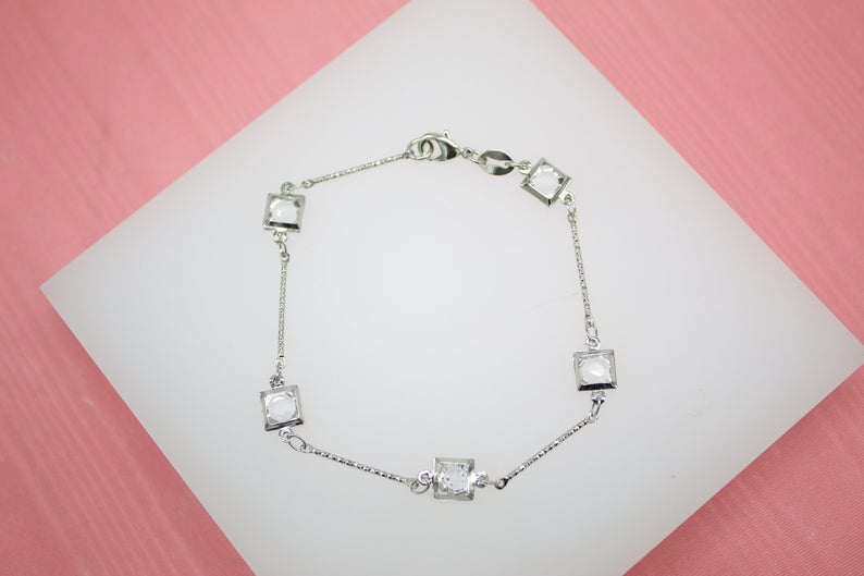 A delicate silver chain bracelet featuring sparkling square-cut white crystals rests gracefully on a white display surface against a pink background.