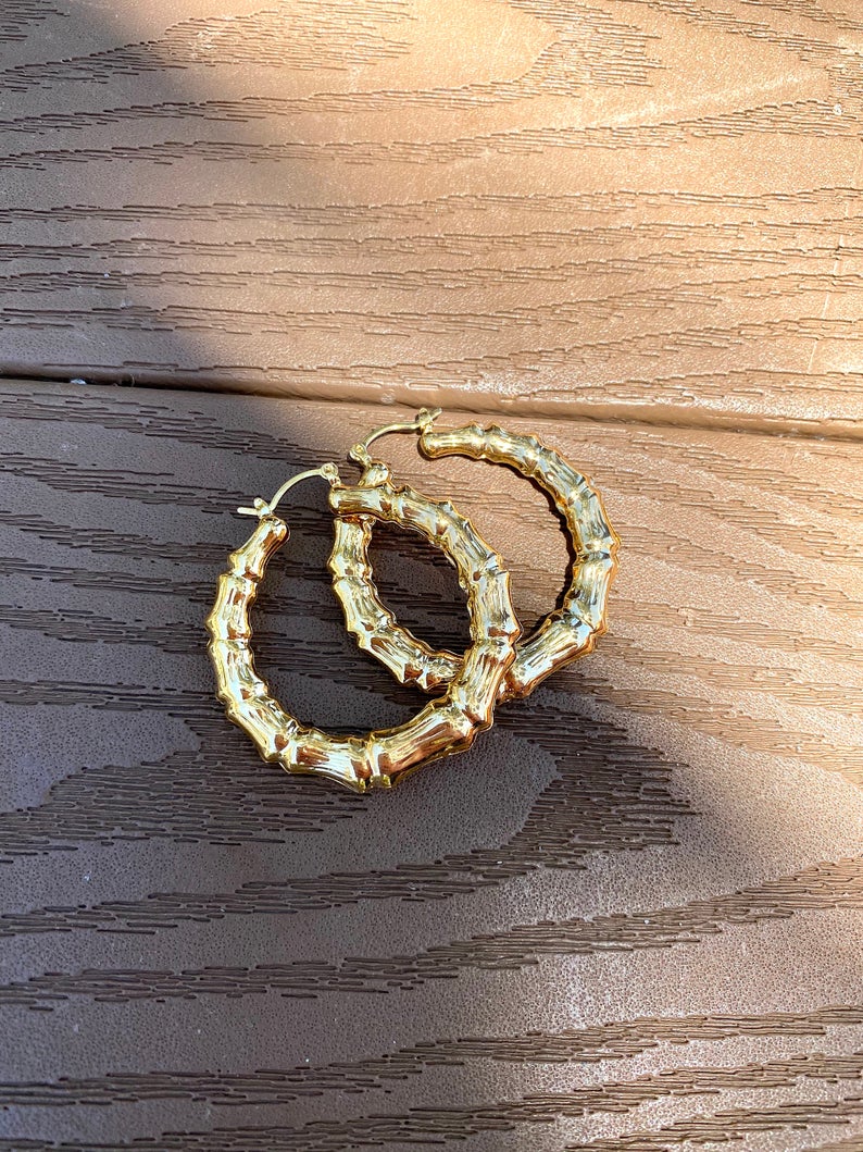A pair of polished gold bamboo hoop earrings rests on a textured wooden deck.