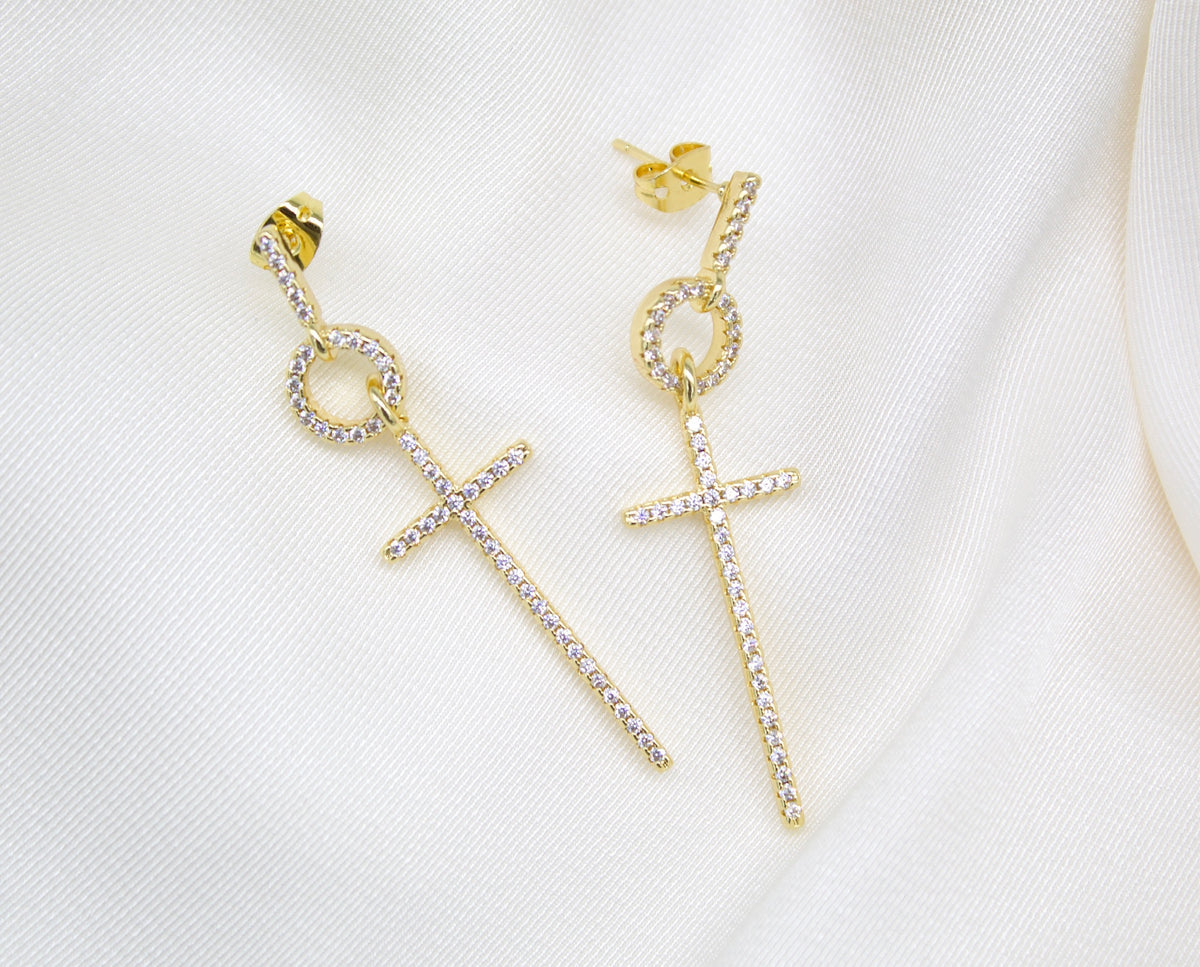 A pair of gold dangle earrings featuring sparkling pavé-set cubic zirconia stones in a cross design rest on a white silk background.