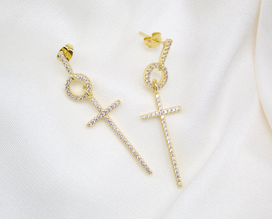 A pair of gold dangle earrings featuring sparkling pavé-set cubic zirconia stones in a cross design rest on a white silk background.