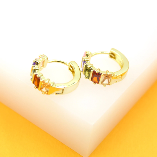 A pair of gold huggie hoop earrings featuring multicolored baguette-cut cubic zirconia stones rests on a white and orange surface.
