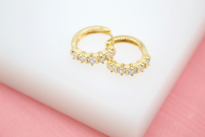 A pair of sparkling gold hoop earrings with small square zirconia stones rests on a white and pink surface.