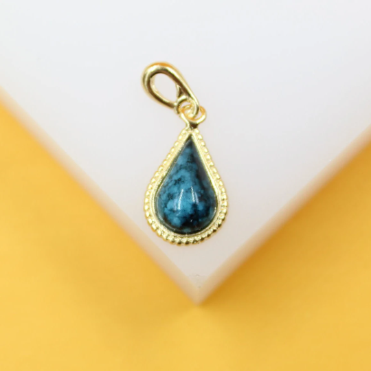 A gold teardrop pendant necklace features a marbled turquoise stone, resting on a white display block against a yellow background.