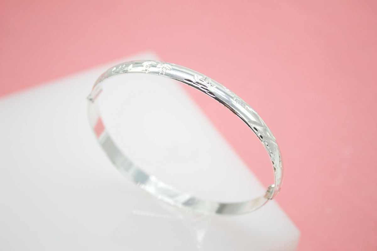 A polished silver baby bangle bracelet rests on a white display surface against a pink background.