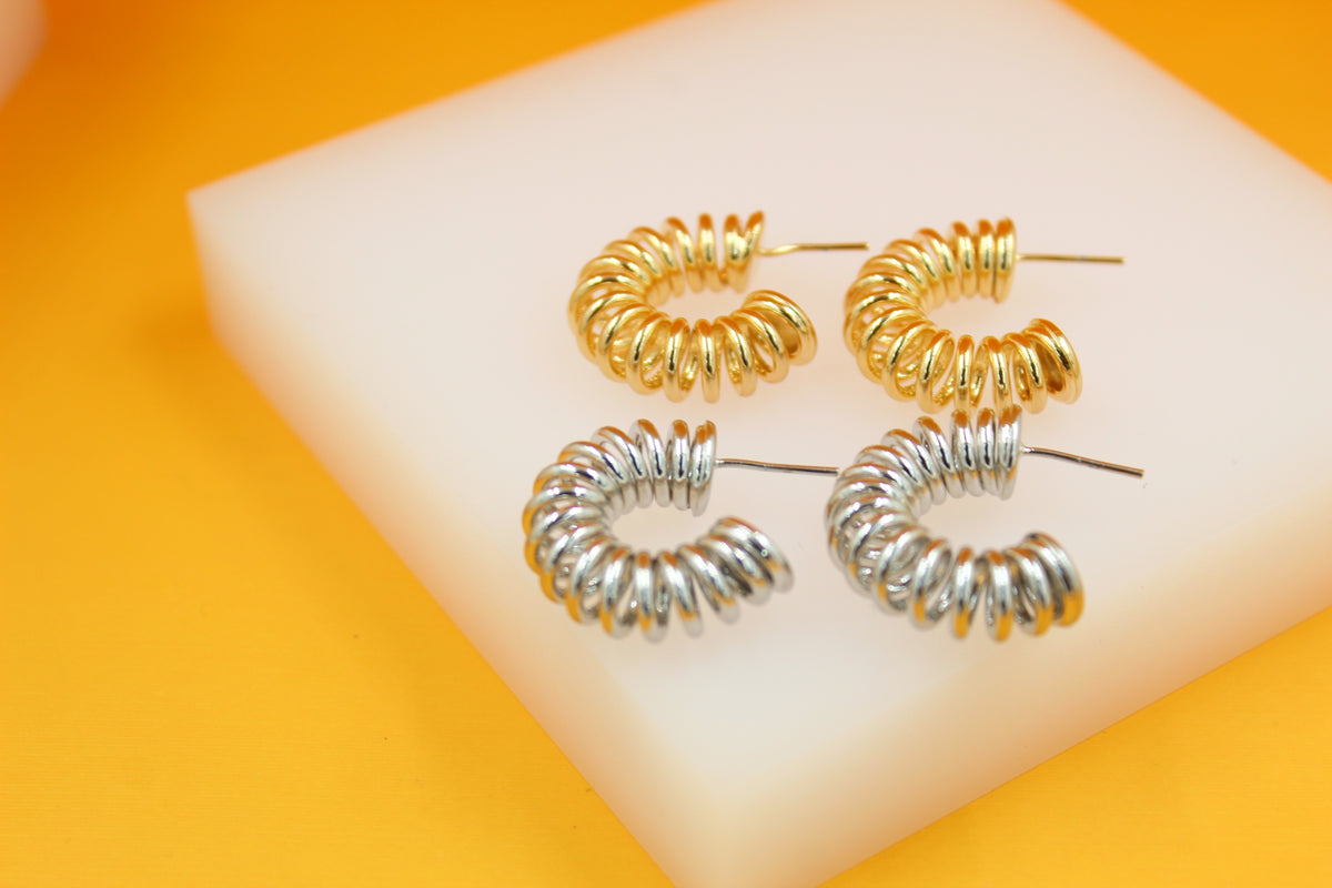 Two pairs of spiral-shaped gold and silver hoop earrings rest neatly on a white display block against a vibrant yellow background.