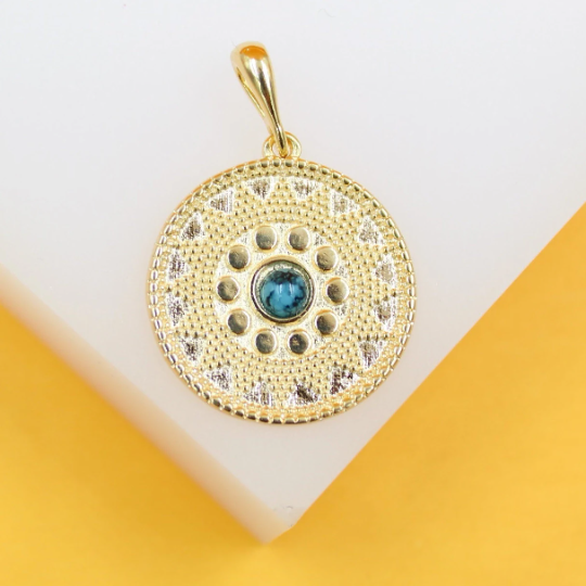 A gold pendant with intricate geometric patterns and a central turquoise stone rests on a white display surface.