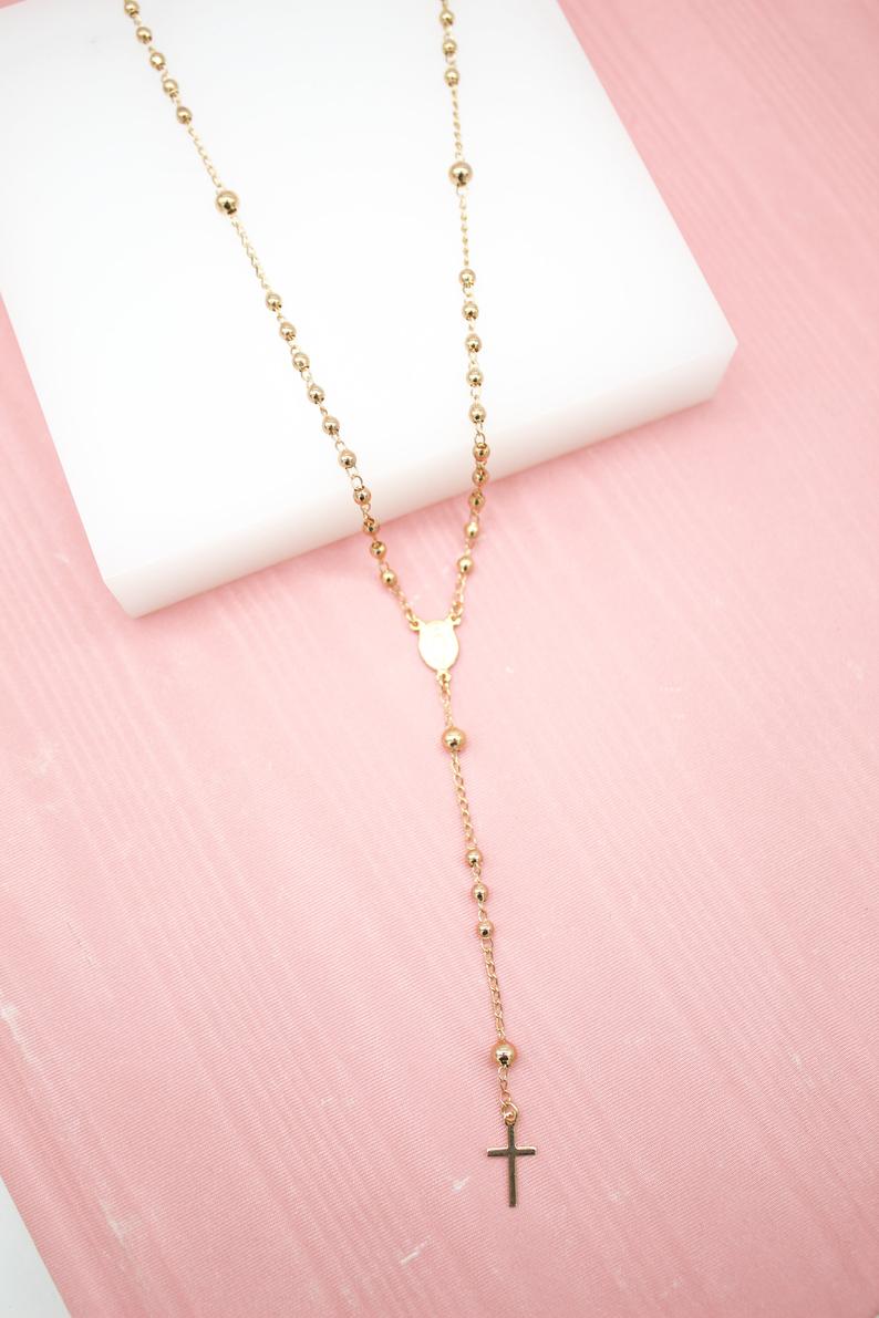 A delicate gold rosary necklace featuring shiny spherical beads rests against a white block on a soft pink background.