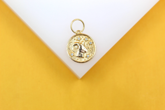 A gold-plated Aries zodiac charm with sparkling CZ stones rests on a white and yellow display surface.