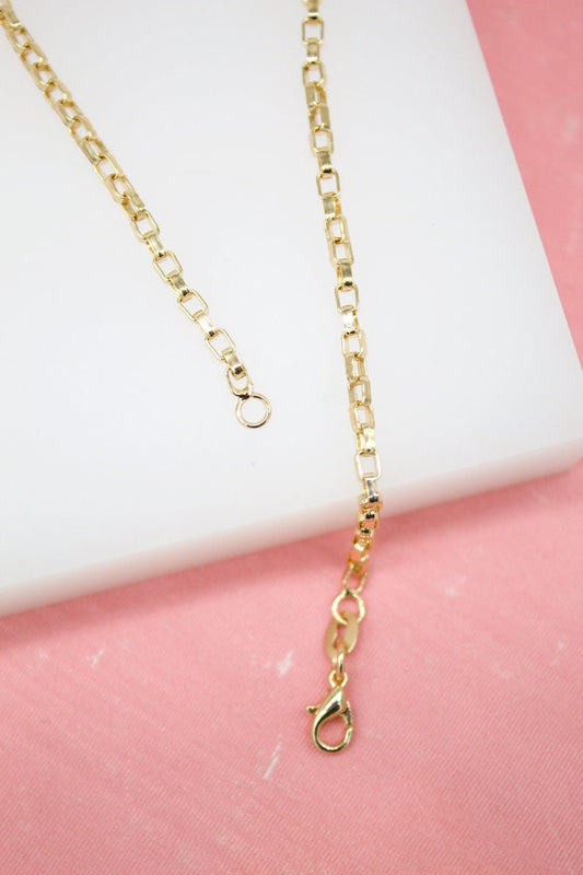 A polished gold box chain necklace rests on a white display against a pink background, showcasing its lobster claw clasp and extender rings.