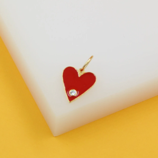 A gold heart-shaped pendant featuring vibrant red enamel and a sparkling cubic zirconia stone rests on a white display surface.