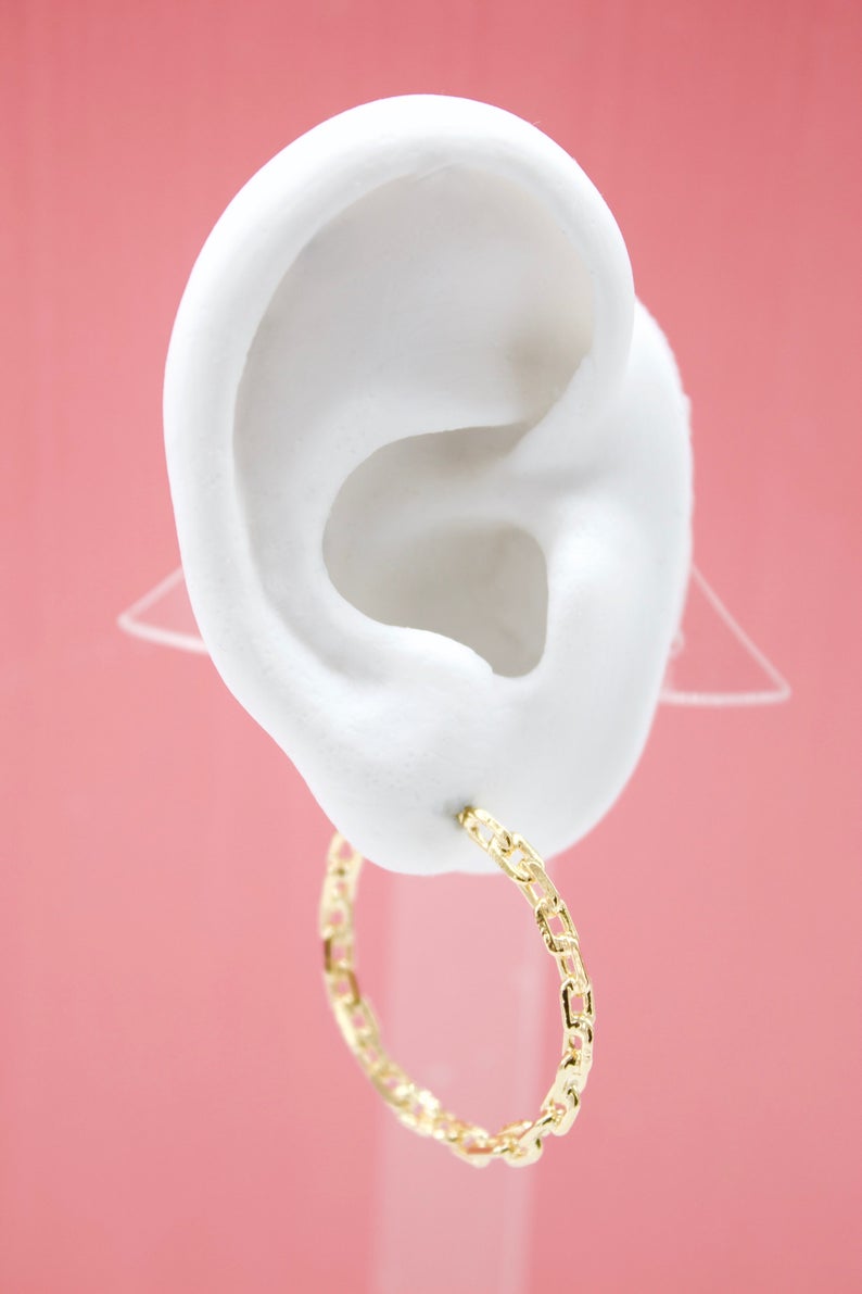 A white mannequin ear displays a textured gold chain-link hoop earring against a solid pink background.