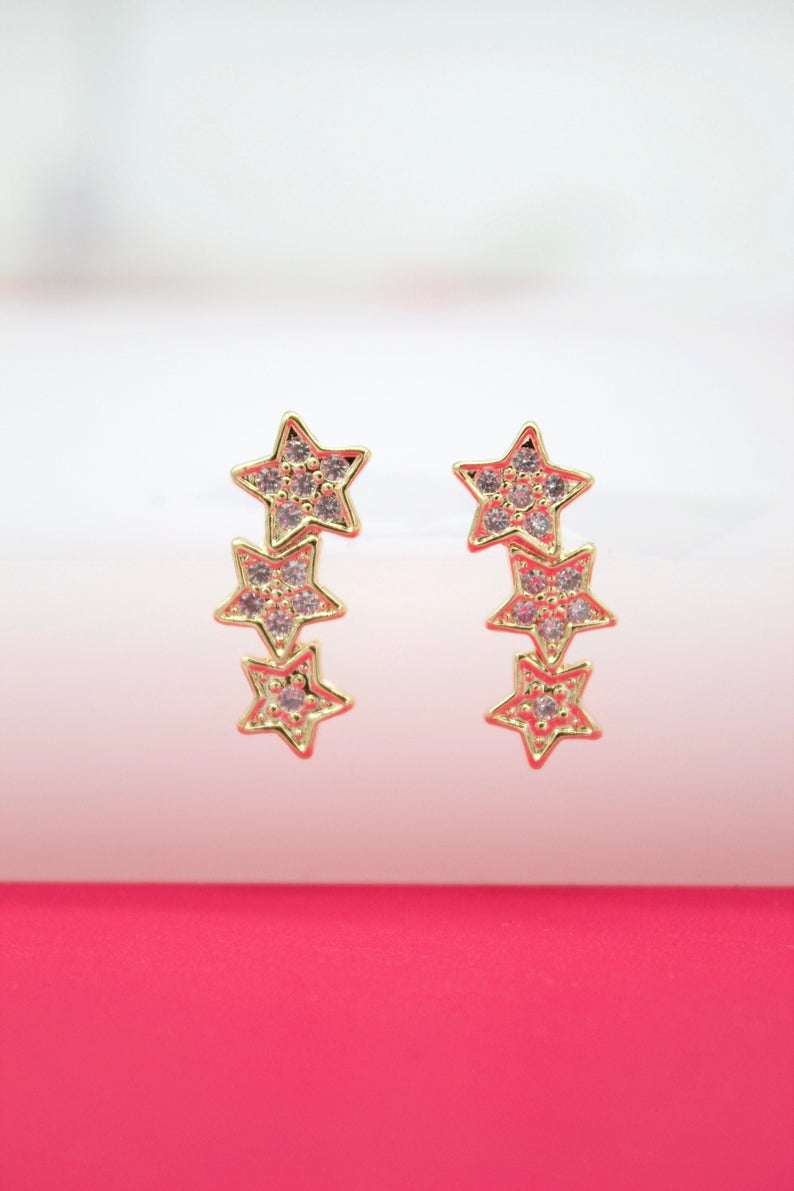 A pair of gold star crawler earrings featuring sparkling pink crystal-encrusted designs rests on a bright pink surface.