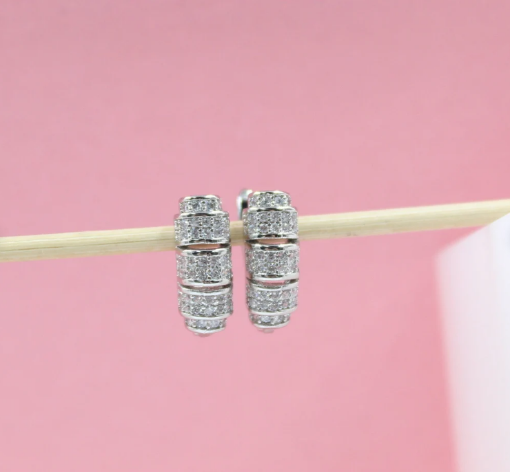 A pair of sparkling paved silver hoop earrings rests on a wooden stick against a soft pink backdrop.