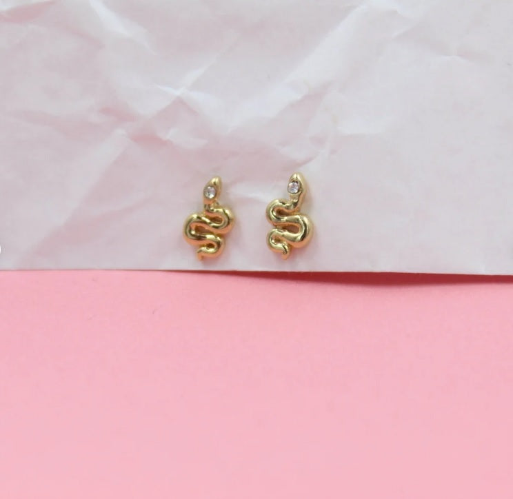A pair of gold snake stud earrings featuring tiny crystal eyes rests against a pink and white backdrop.