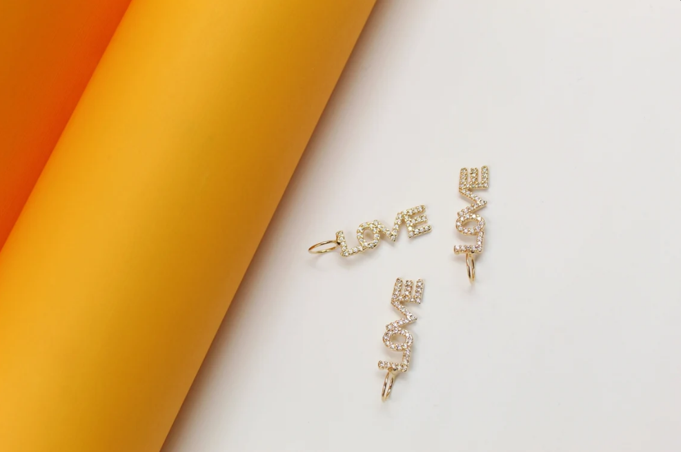Three gold "LOVE" charms embellished with sparkling cubic zirconia crystals rest against a white surface next to a bright yellow and orange cylindrical backdrop.