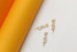Three gold "LOVE" charms embellished with sparkling cubic zirconia crystals rest against a white surface next to a bright yellow and orange cylindrical backdrop.