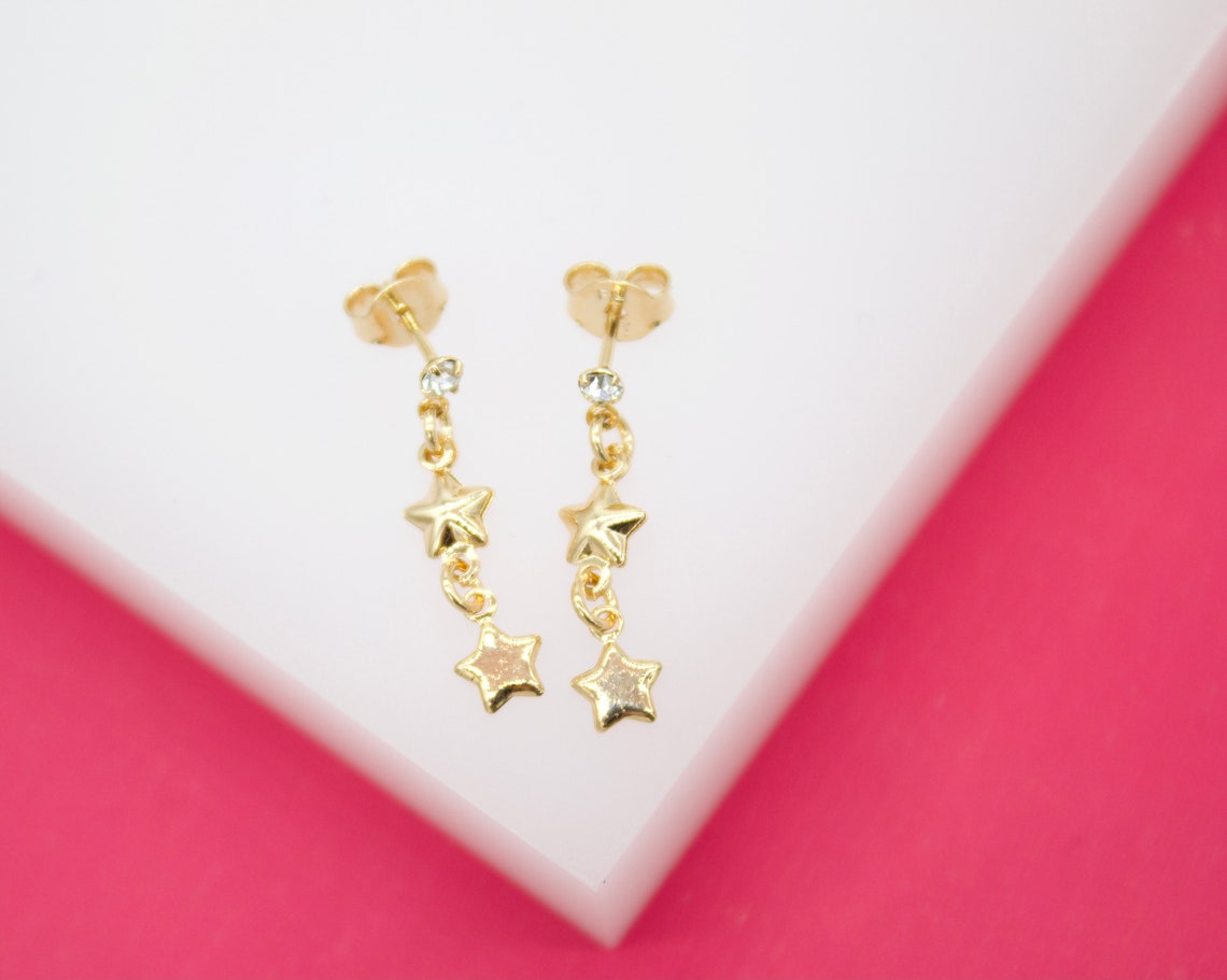 Two delicate gold star drop earrings rest on a white surface against a vibrant pink background.