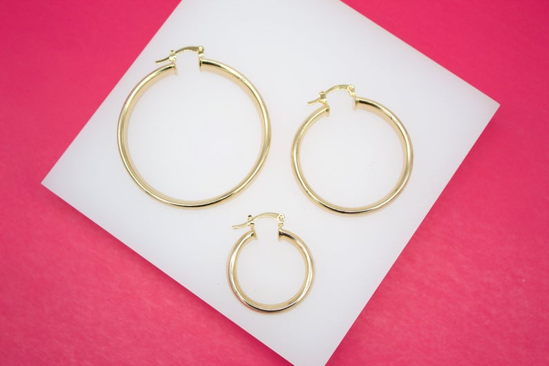 Three graduated sizes of polished gold hoop earrings lie arranged on a white display surface against a vibrant pink background.