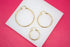Three graduated sizes of polished gold hoop earrings lie arranged on a white display surface against a vibrant pink background.