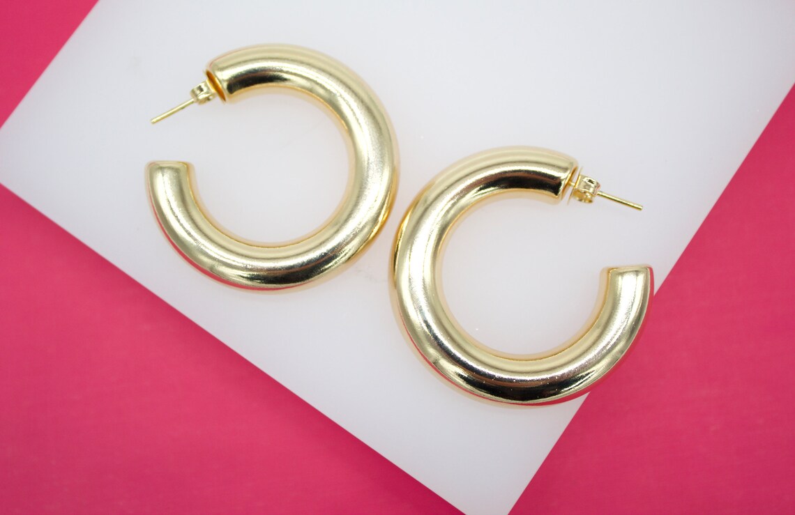 A pair of thick, polished gold hoop earrings rests on a white and bright pink display surface.