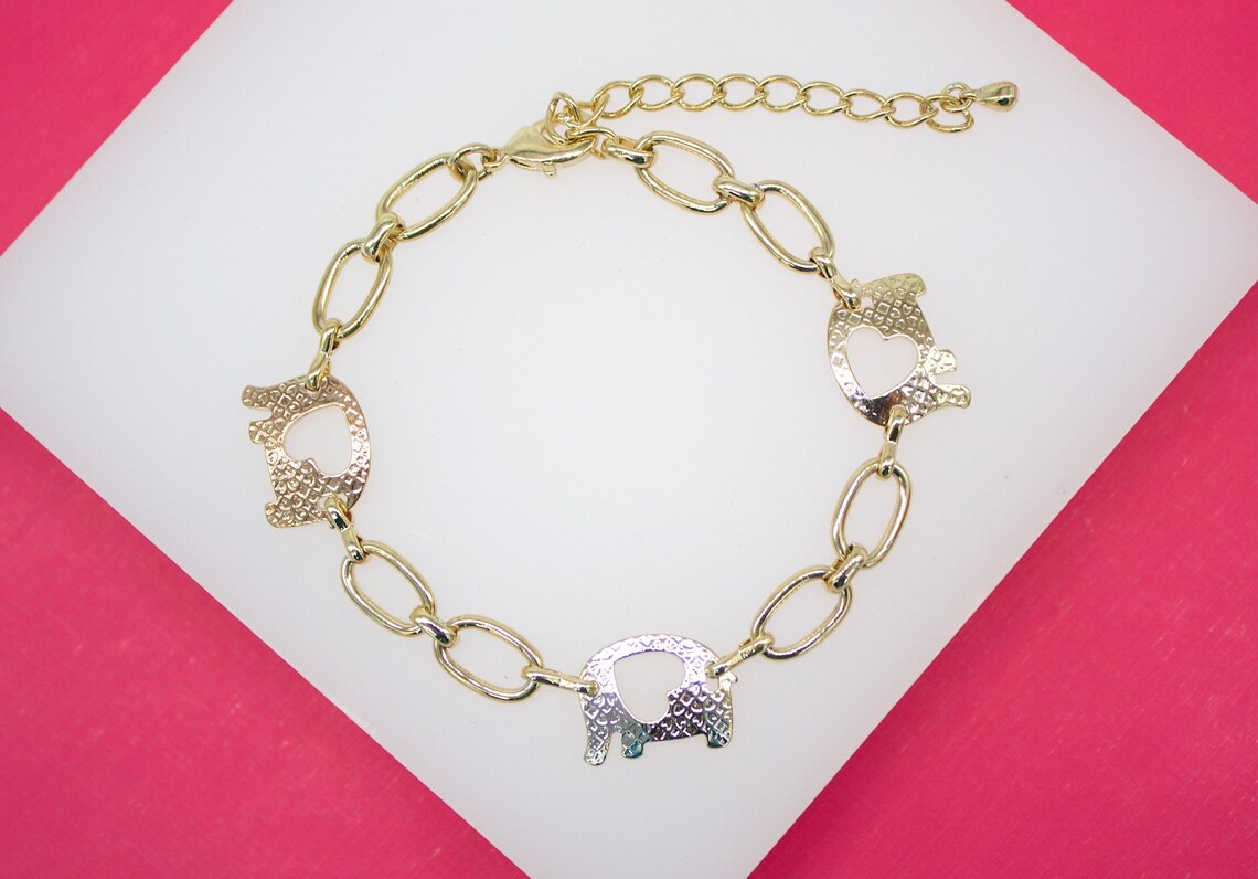 A gold-plated chain bracelet featuring three elephant charms with cutout hearts lies on a white display surface against a vibrant pink background.