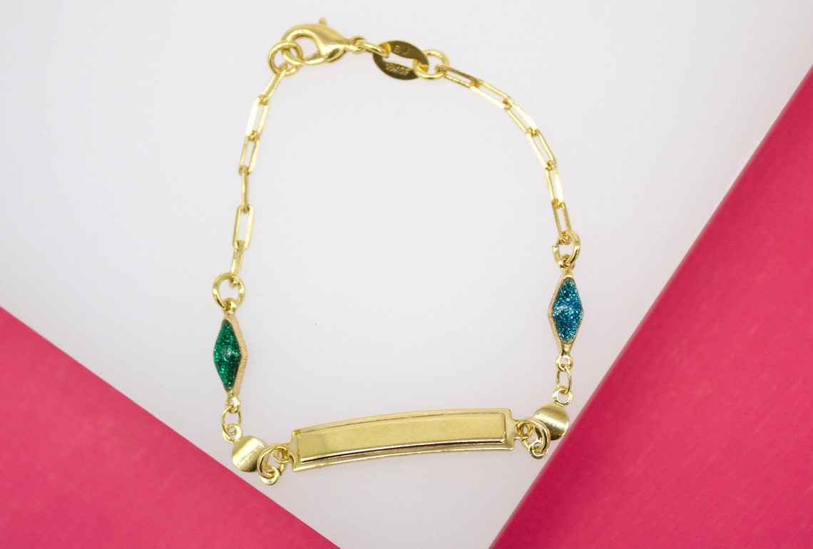 A gold ID bracelet features a link chain adorned with a green and a blue glitter diamond-shaped charm.