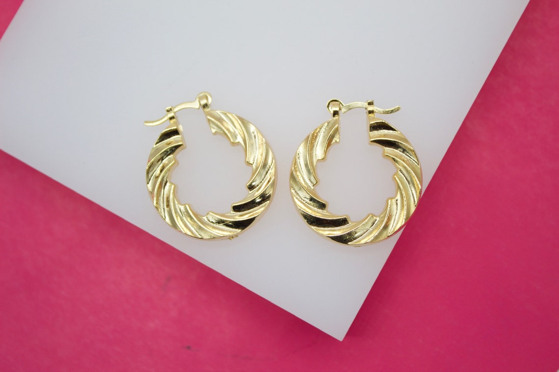 Two gold twist hoop earrings rest on a white surface against a bright pink background.