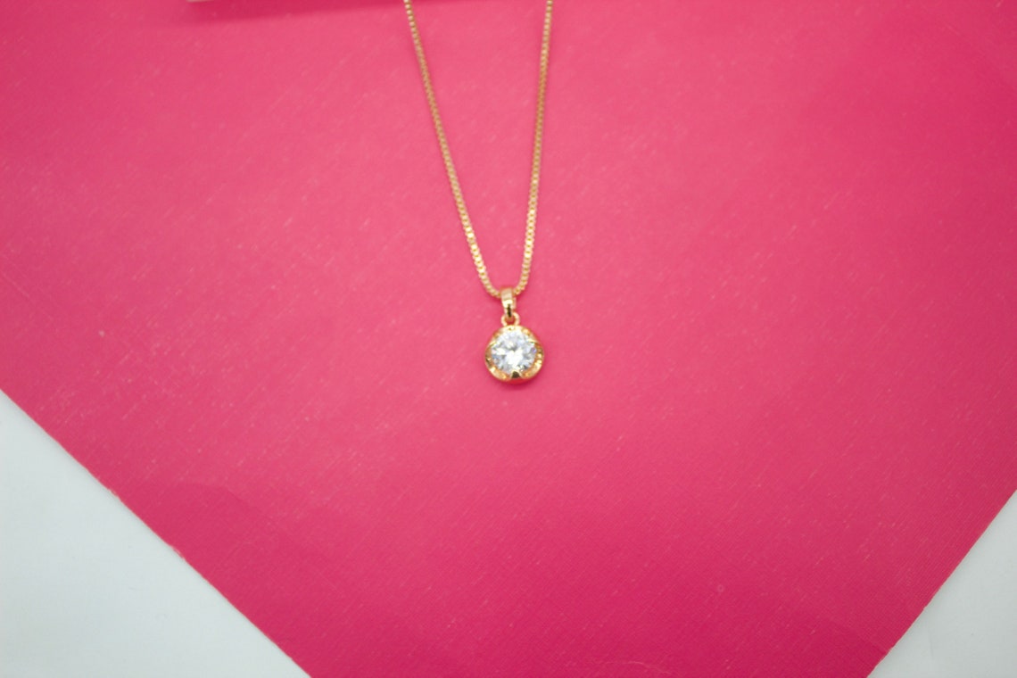A delicate gold chain necklace with a round solitaire diamond pendant rests against a bright pink background.