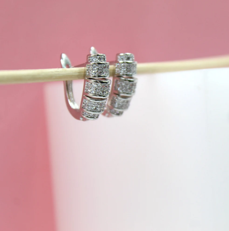 Two silver hoop earrings encrusted with sparkling cubic zirconia stones hang from a thin wooden stick against a pink and white background.