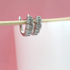 Two silver hoop earrings encrusted with sparkling cubic zirconia stones hang from a thin wooden stick against a pink and white background.