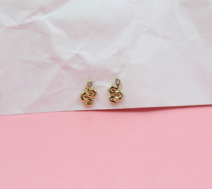 Two gold snake stud earrings with crystal eyes rest on a pink and white textured surface.