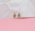Two gold snake stud earrings with crystal eyes rest on a pink and white textured surface.
