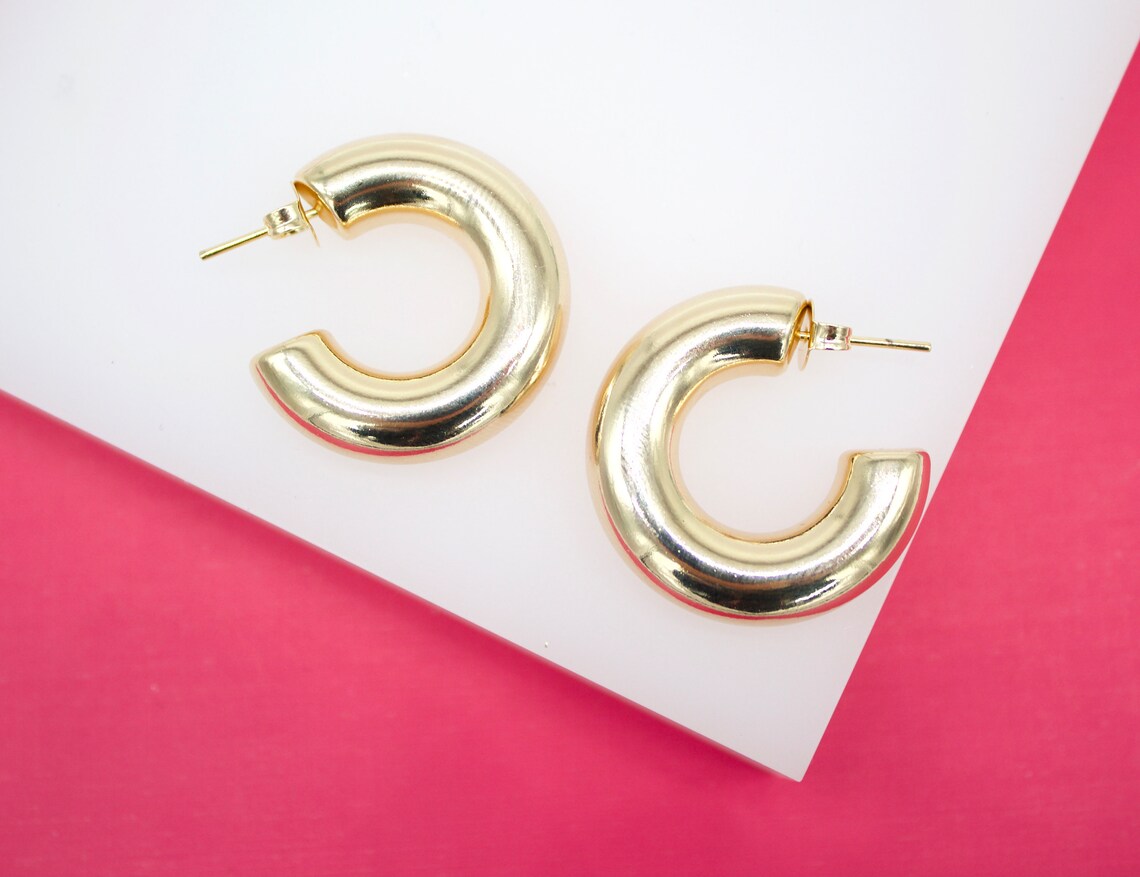 A pair of thick, polished gold hoop earrings rests on a white and bright pink display block.