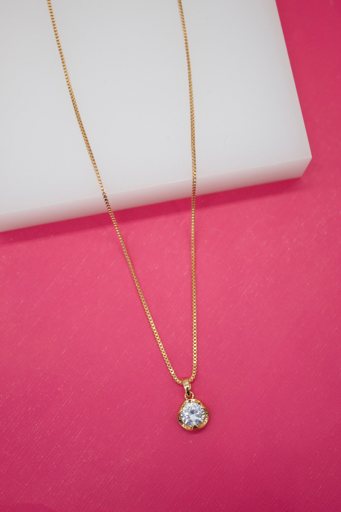 A delicate gold box chain necklace with a sparkling solitaire diamond pendant rests against a vibrant pink and white backdrop.