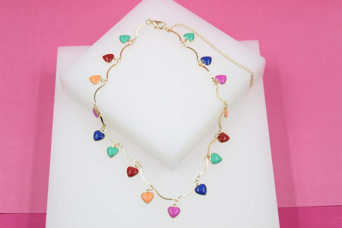 A gold-tone charm necklace featuring colorful enamel hearts displayed on a white platform against a pink background.