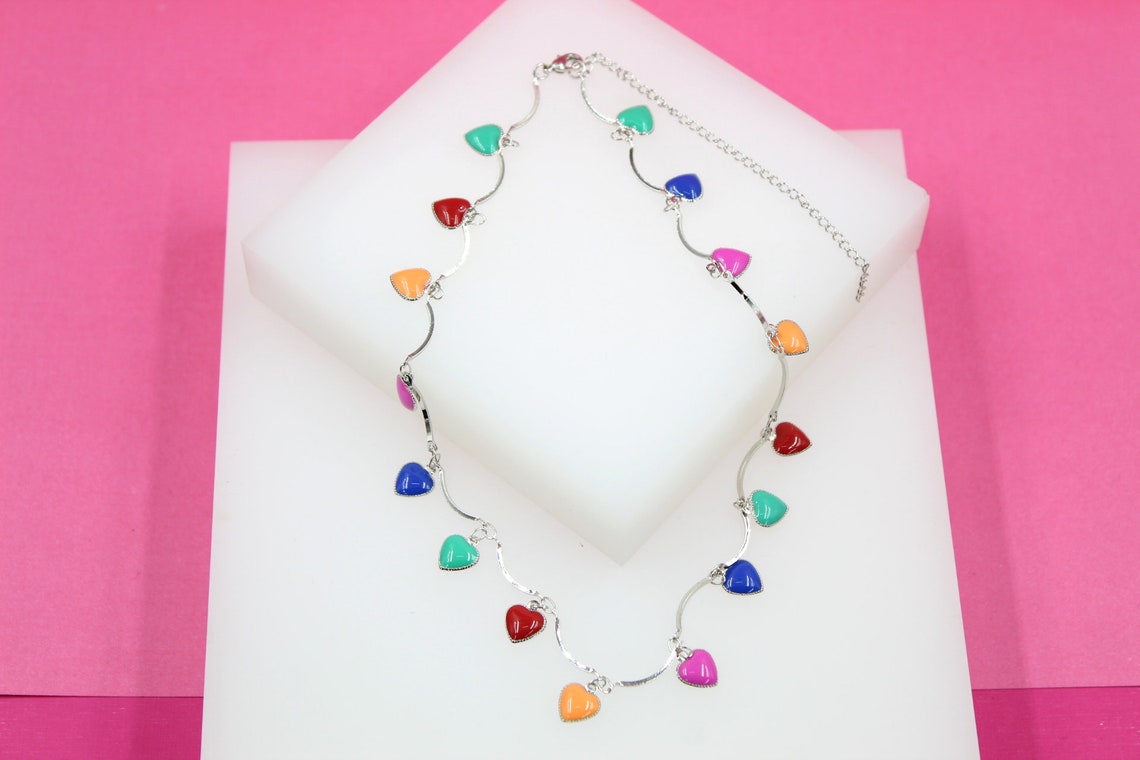 A silver necklace adorned with multicolored enamel heart charms rests on a white display block against a vibrant pink background.