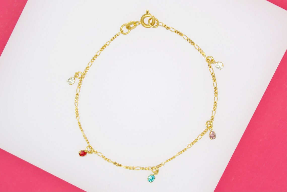 A delicate gold chain bracelet features colorful hanging crystal charms against a pink and white background.